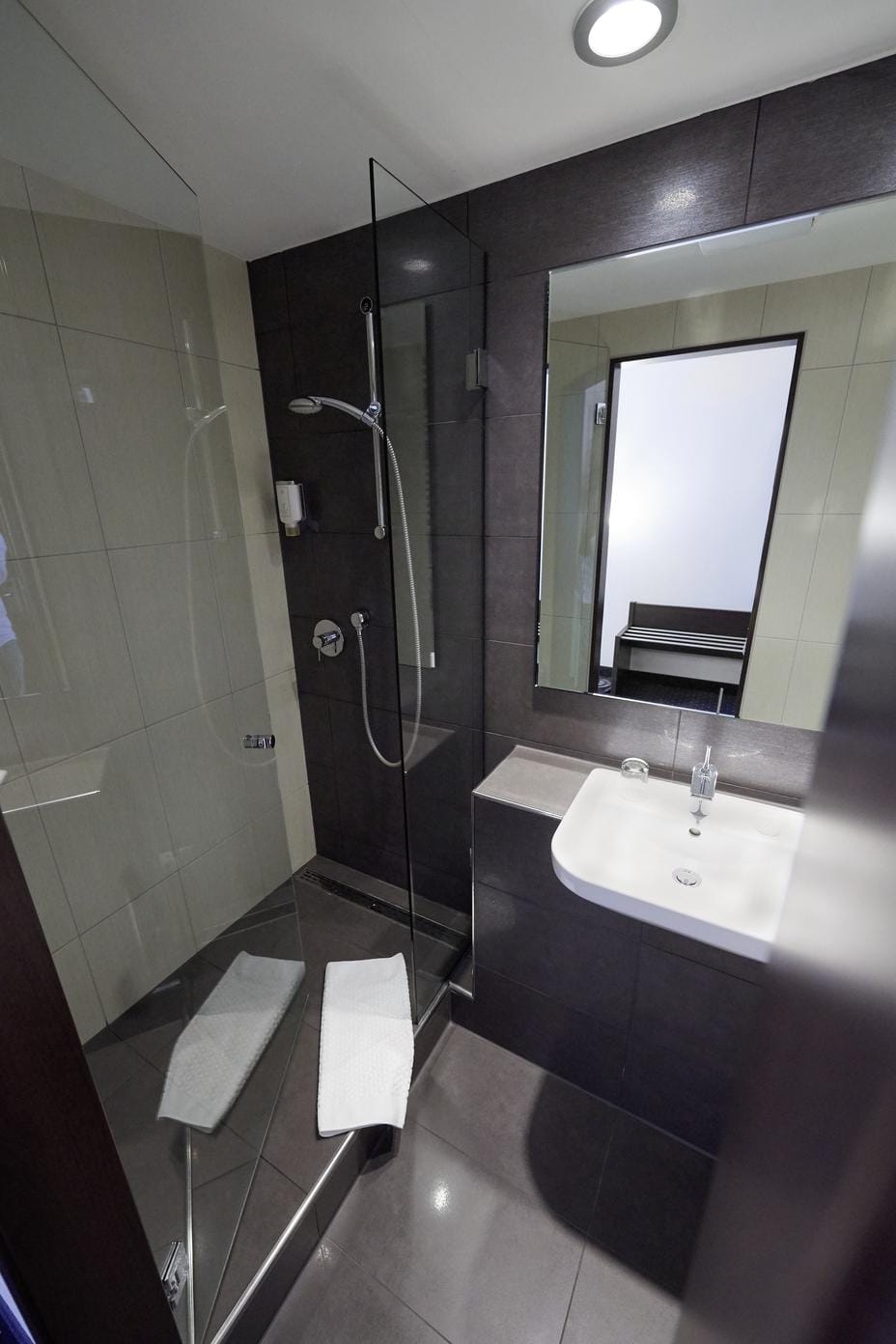 Modernes Hotelbad mit dunklen Fliesen, Glasdusche und weißem Waschbecken.