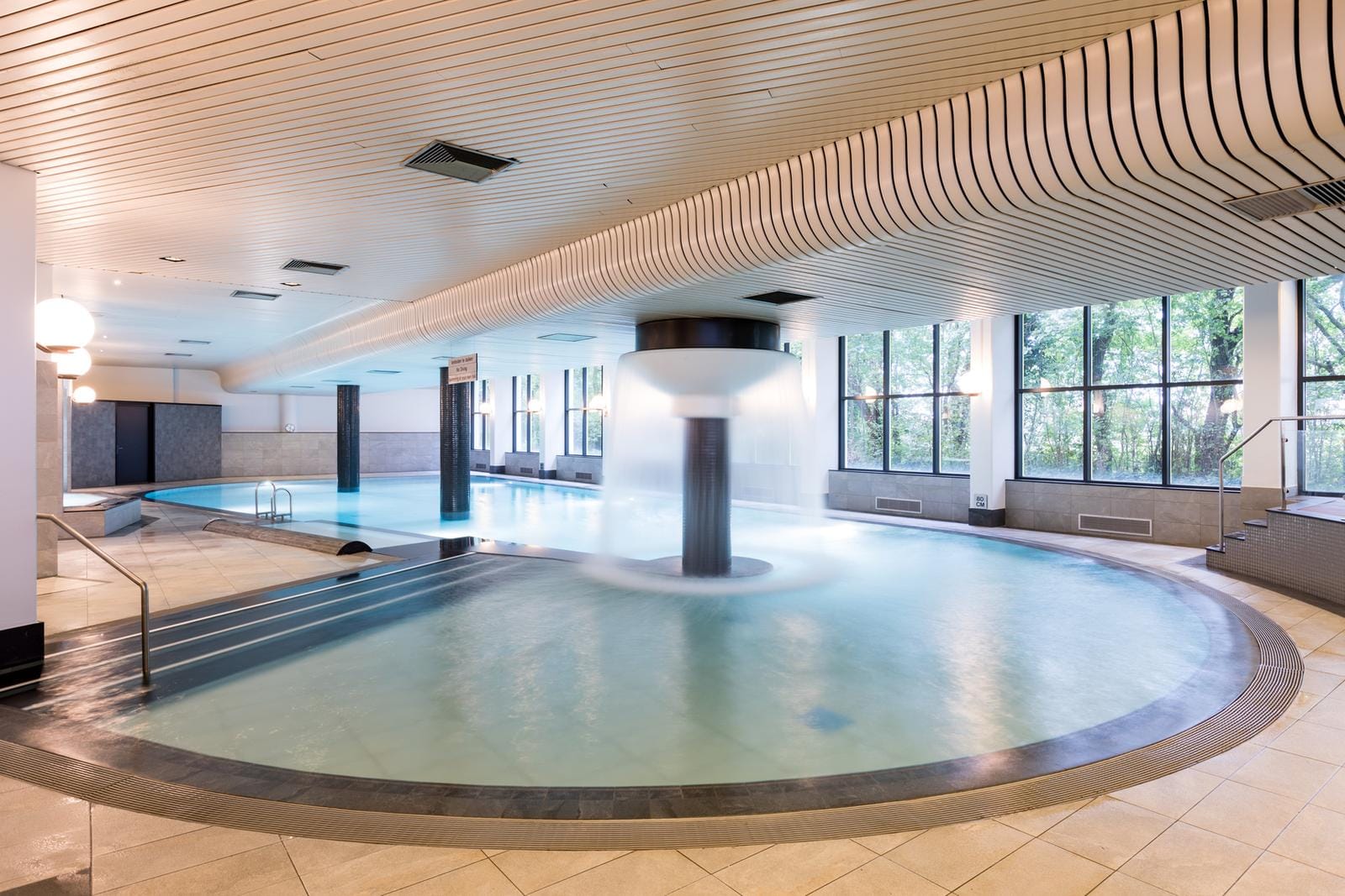 Modernes, helles Spa mit großem Indoor-Pool und Wassersäule, Blick ins Grüne.
