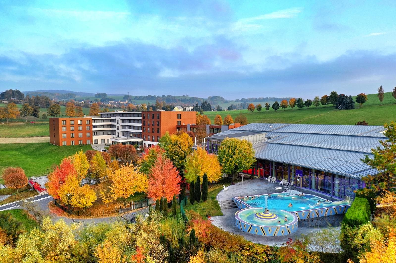 Hotelanlage in herbstlicher Landschaft mit Pool, bunten Bäumen und modernem Gebäude.