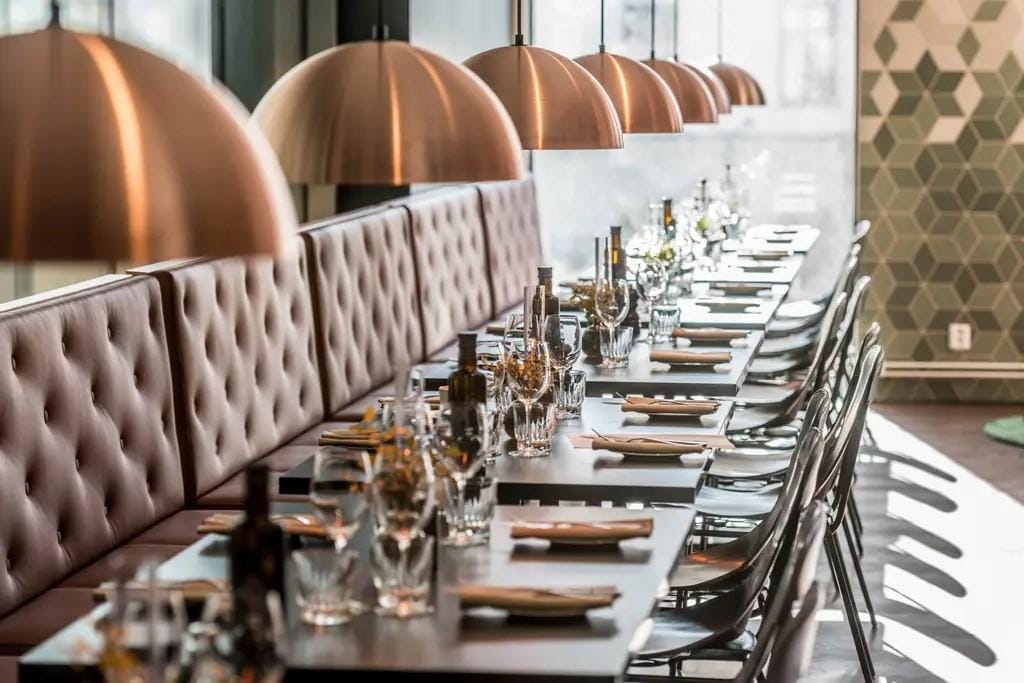 Modernes Hotel-Restaurant mit langen Tischen, Kupferleuchten und gepolsterten Sitzen.