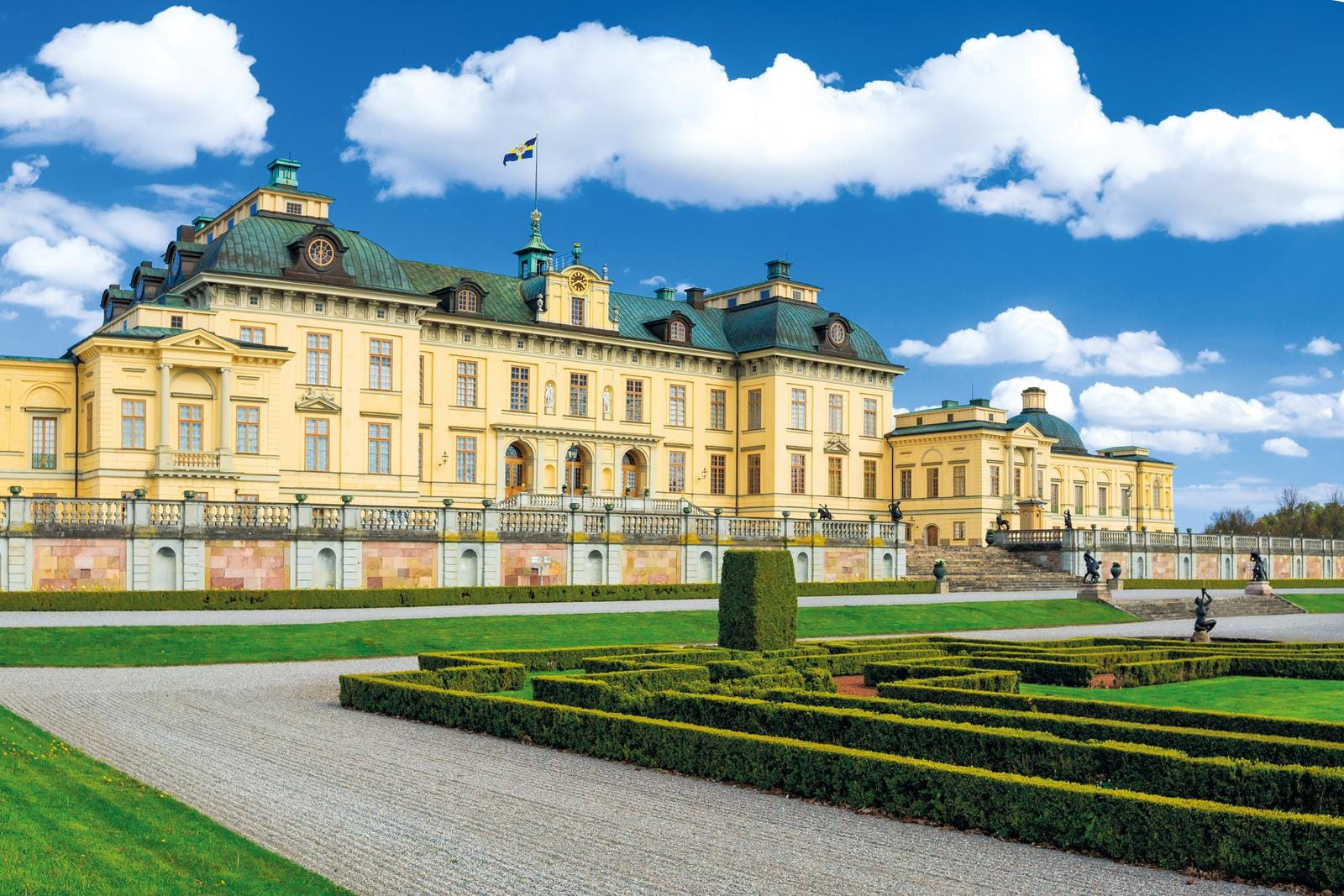 Gelbes Barockschloss mit grünem Dach in gepflegtem Garten, blauer Himmel.