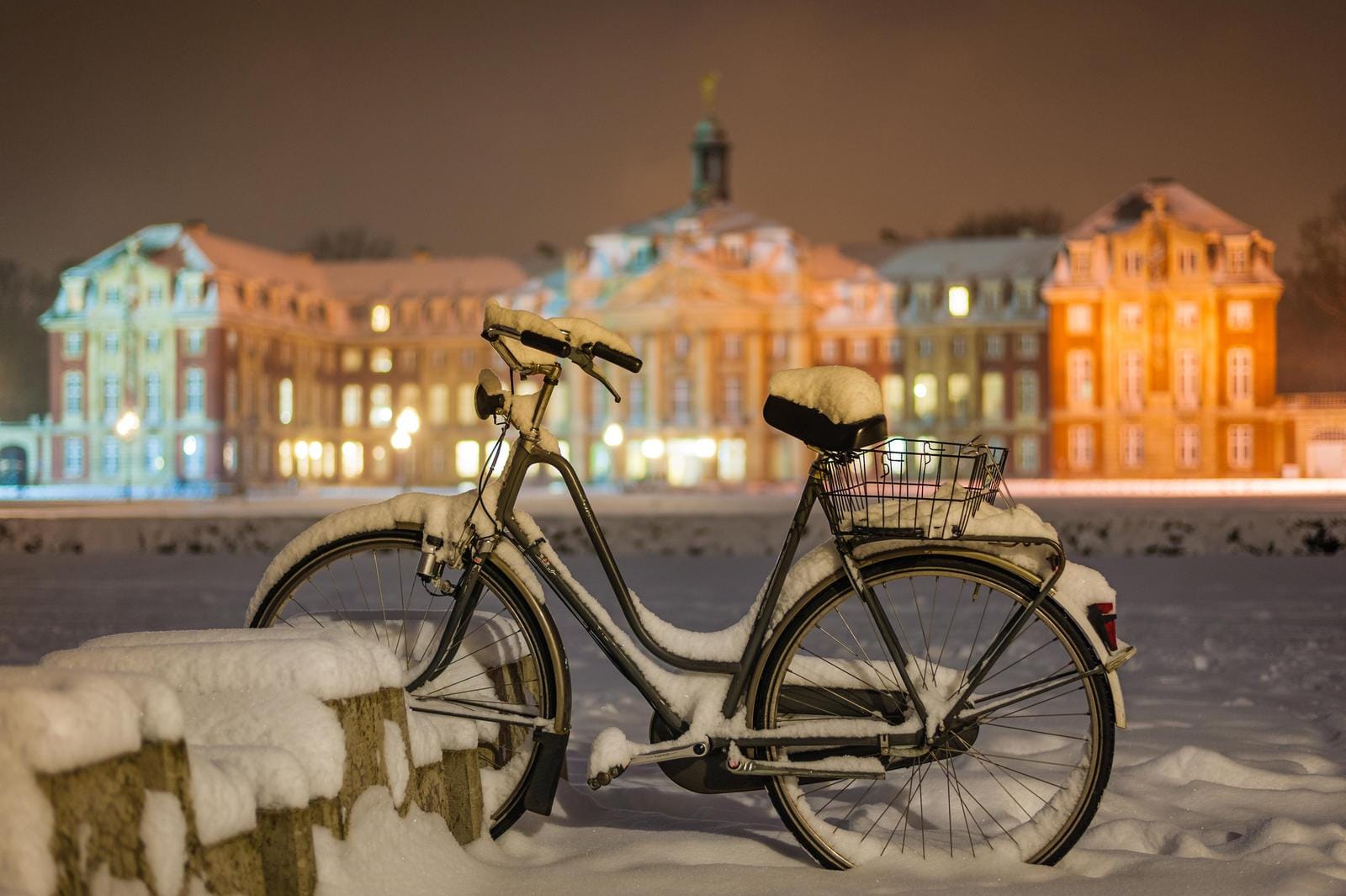 Verschneites Fahrrad vor beleuchtetem Barockschloss in winterlicher Stadtlandschaft.