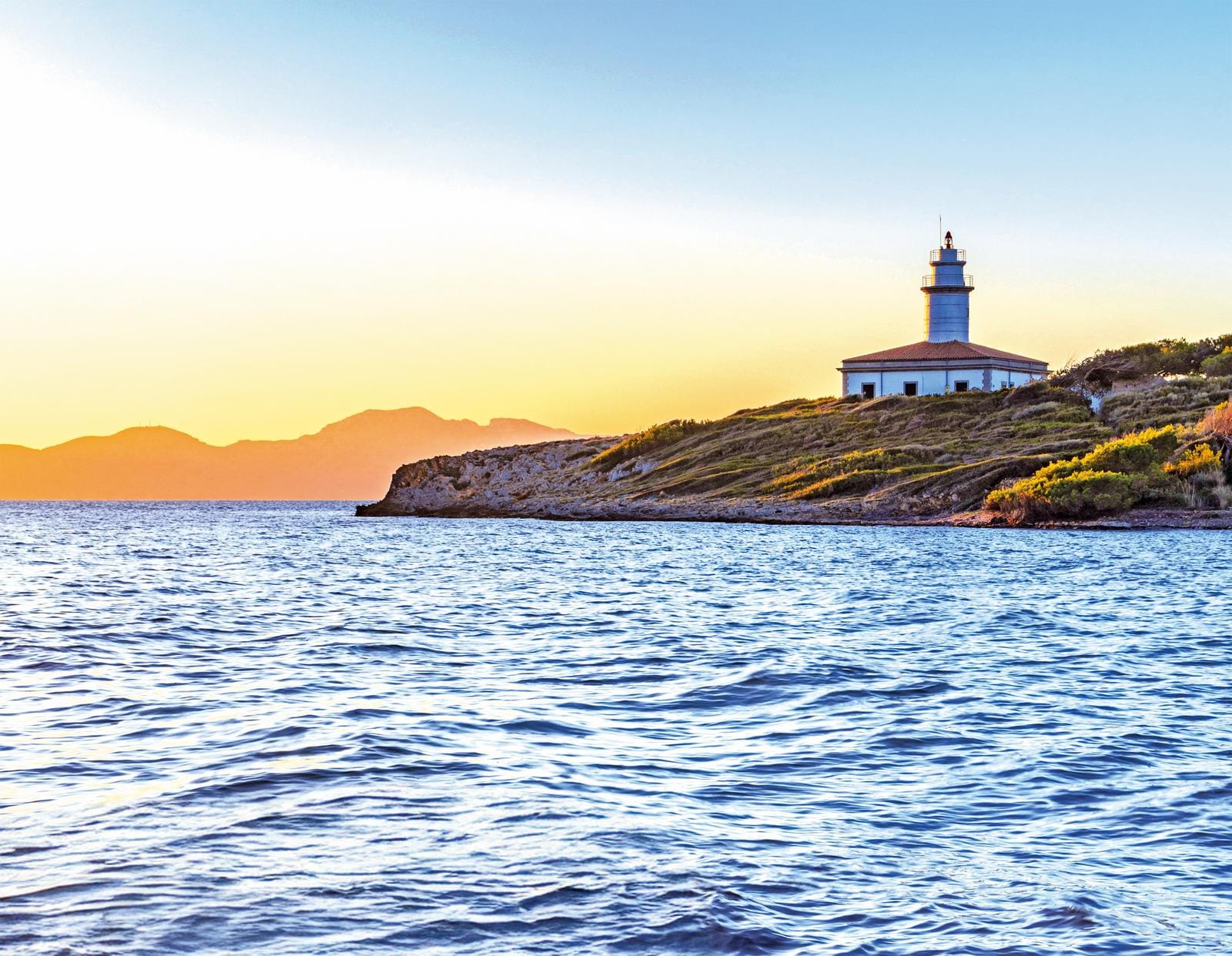 Leuchtturm auf Mallorca bei Sonnenuntergang, umgeben von ruhigem Meer und sanften Hügeln.