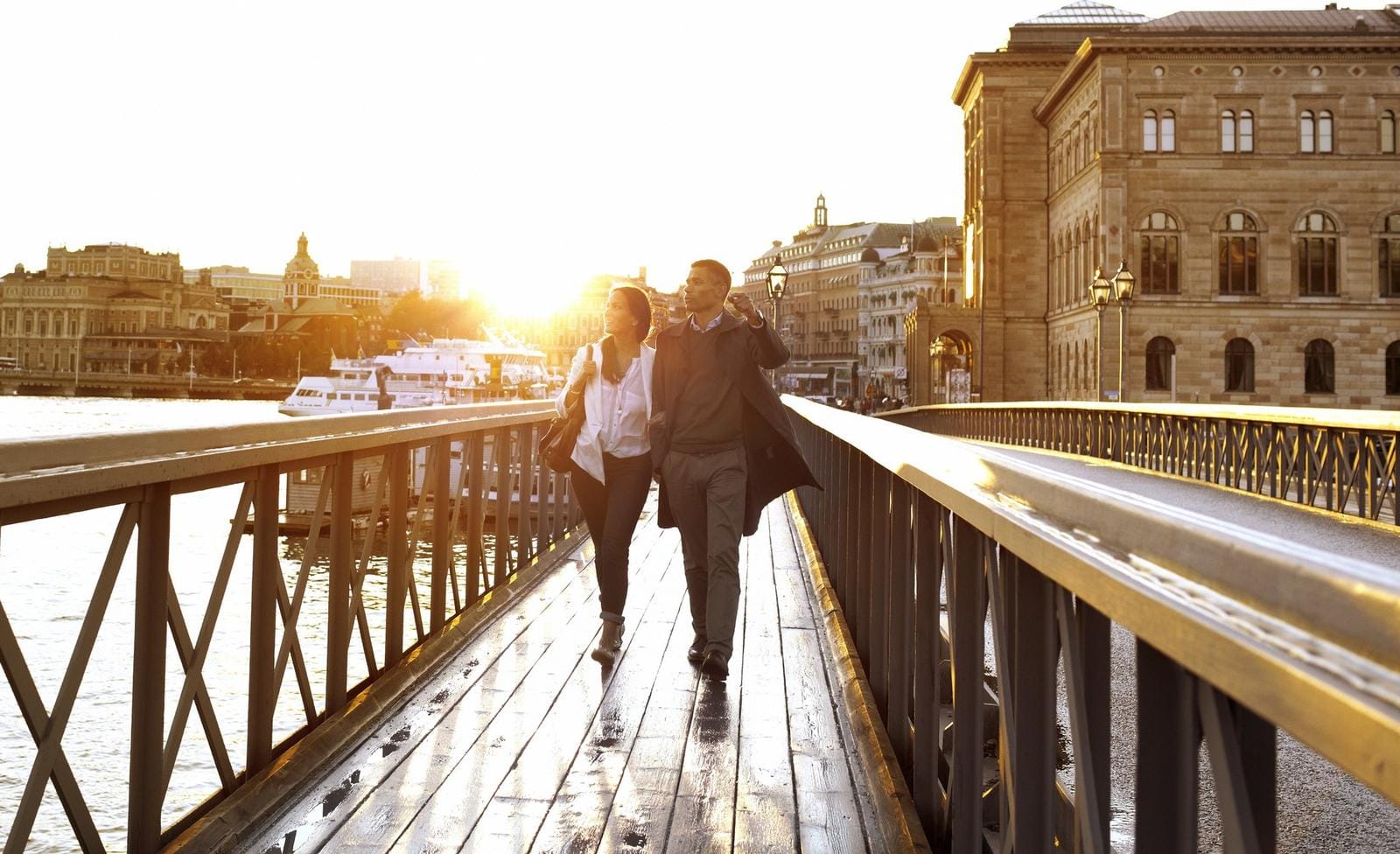 Paar bei Sonnenuntergang auf Brücke, Stadthäuser und Schiffe im Hintergrund, warmes Licht.