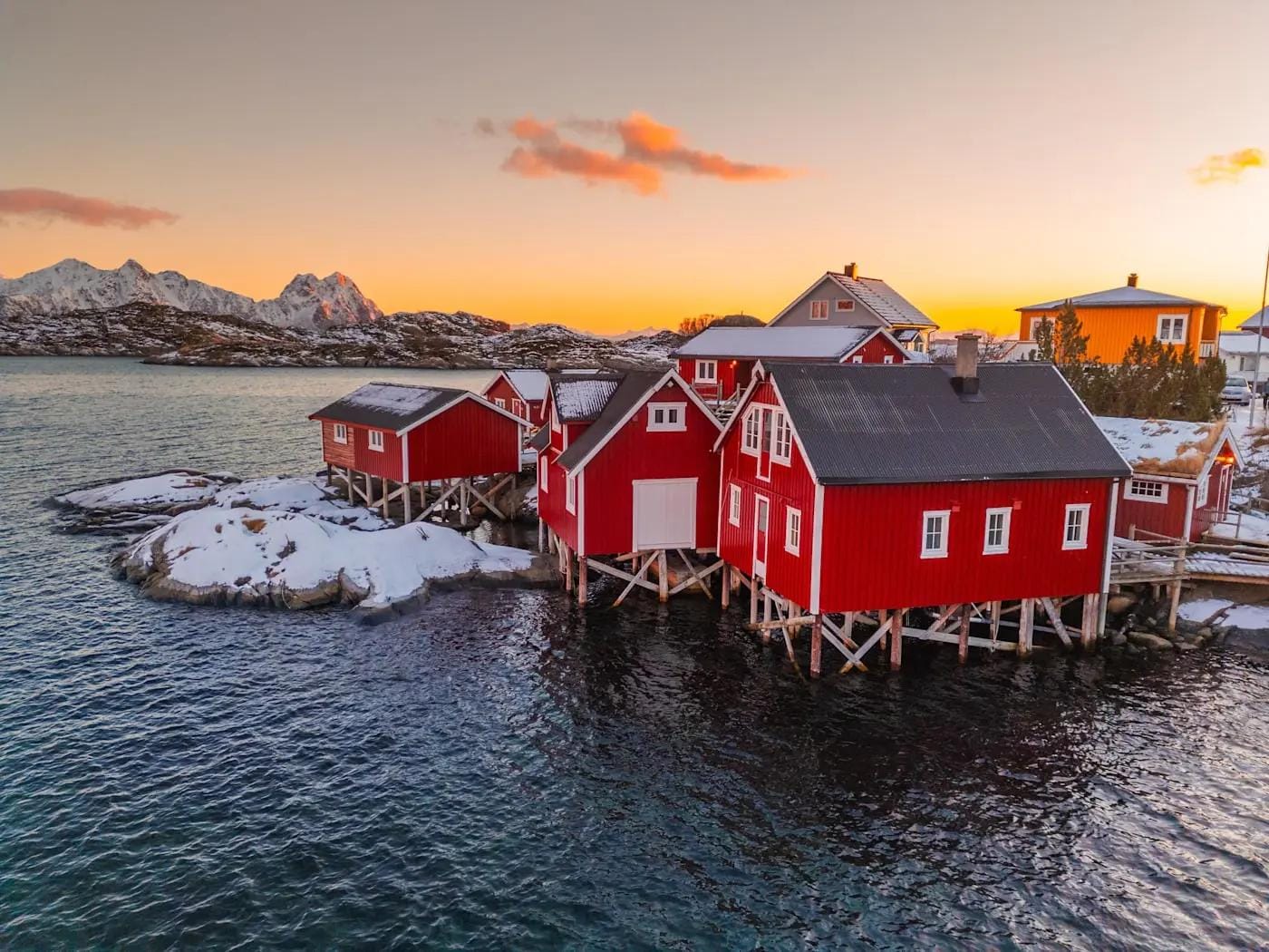 Rote Fischerhütten am Wasser bei Sonnenuntergang, schneebedeckte Berge im Hintergrund.