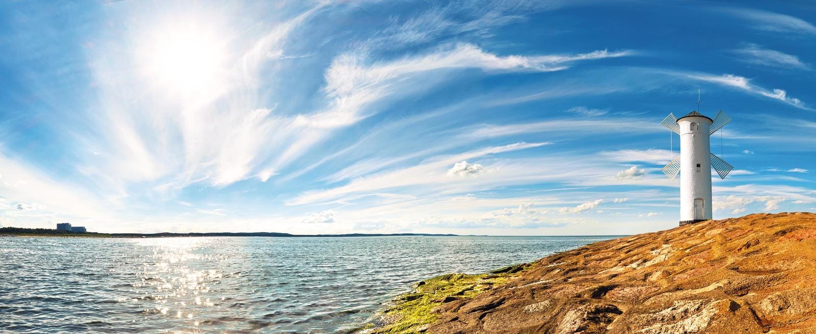Weißer Leuchtturm auf felsiger Küste, blauer Himmel und glitzerndes Meer.