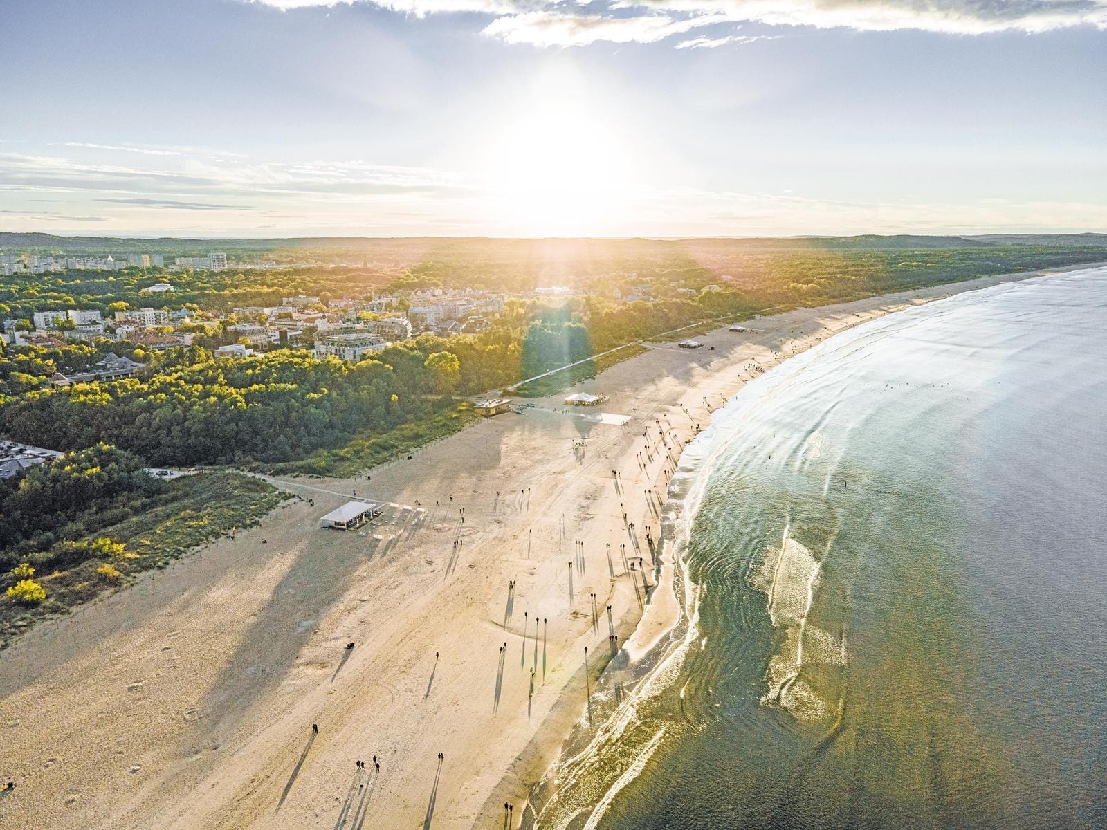 Weitläufiger Strand bei Sonnenuntergang mit sanften Wellen und grüner Stadt im Hintergrund.