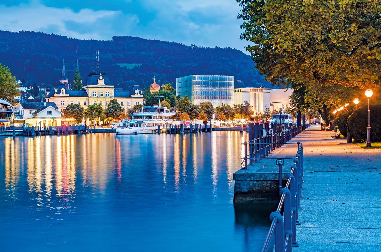 Promenade am Bodensee in Bregenz, Österreich bei Abenddämmerung, mit beleuchteten Gebäuden.