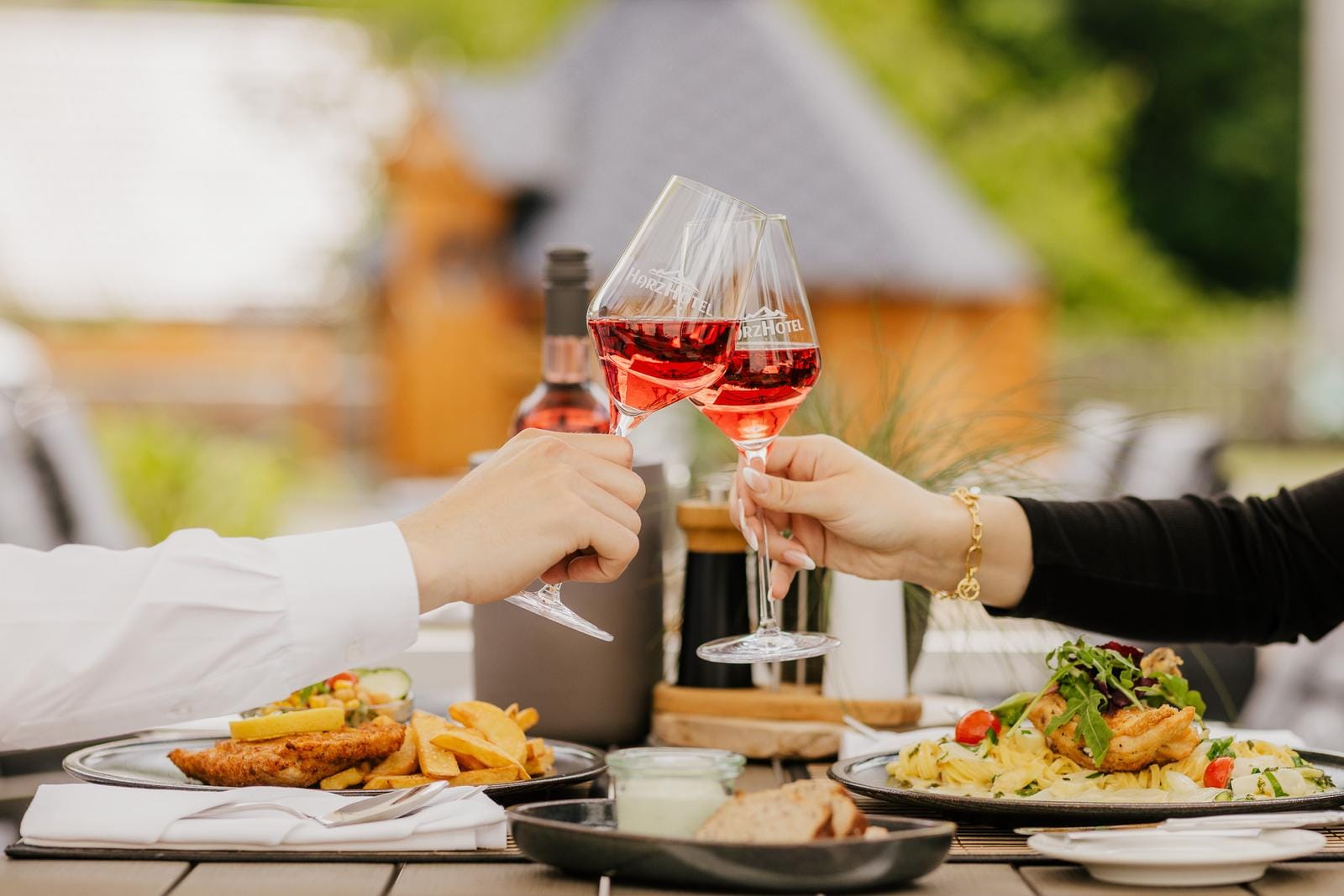 Zwei Hände stoßen mit Roséwein-Gläsern über einem reichhaltigen Essen an.