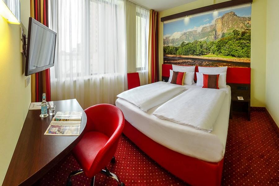 Modernes Hotelzimmer mit Doppelbett, rotem Teppich, Landschaftspanorama und hellen VorhÀngen.