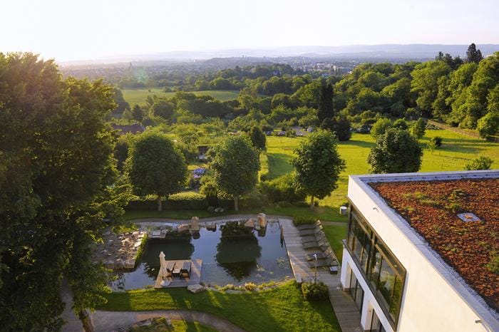 Weitläufige Landschaft mit grünem Hotelgarten, Teich und modernem Gebäude im Sonnenlicht.