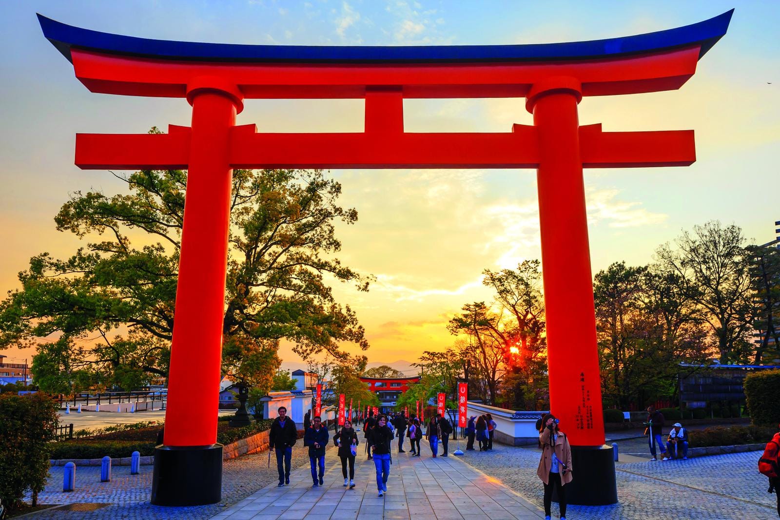 Rotes Torii-Tor in Japan bei Sonnenuntergang, umgeben von Bäumen und Menschen.