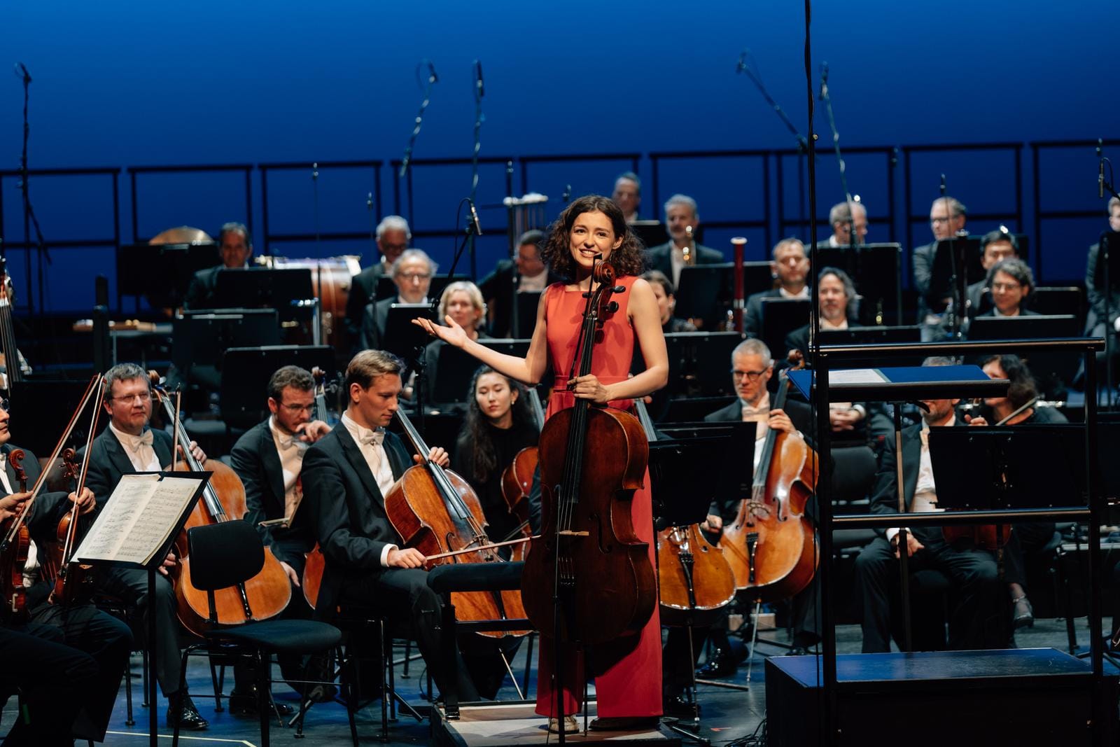 Musikerin im roten Kleid, Orchester im Hintergrund auf Bühne vor blauem Hintergrund.