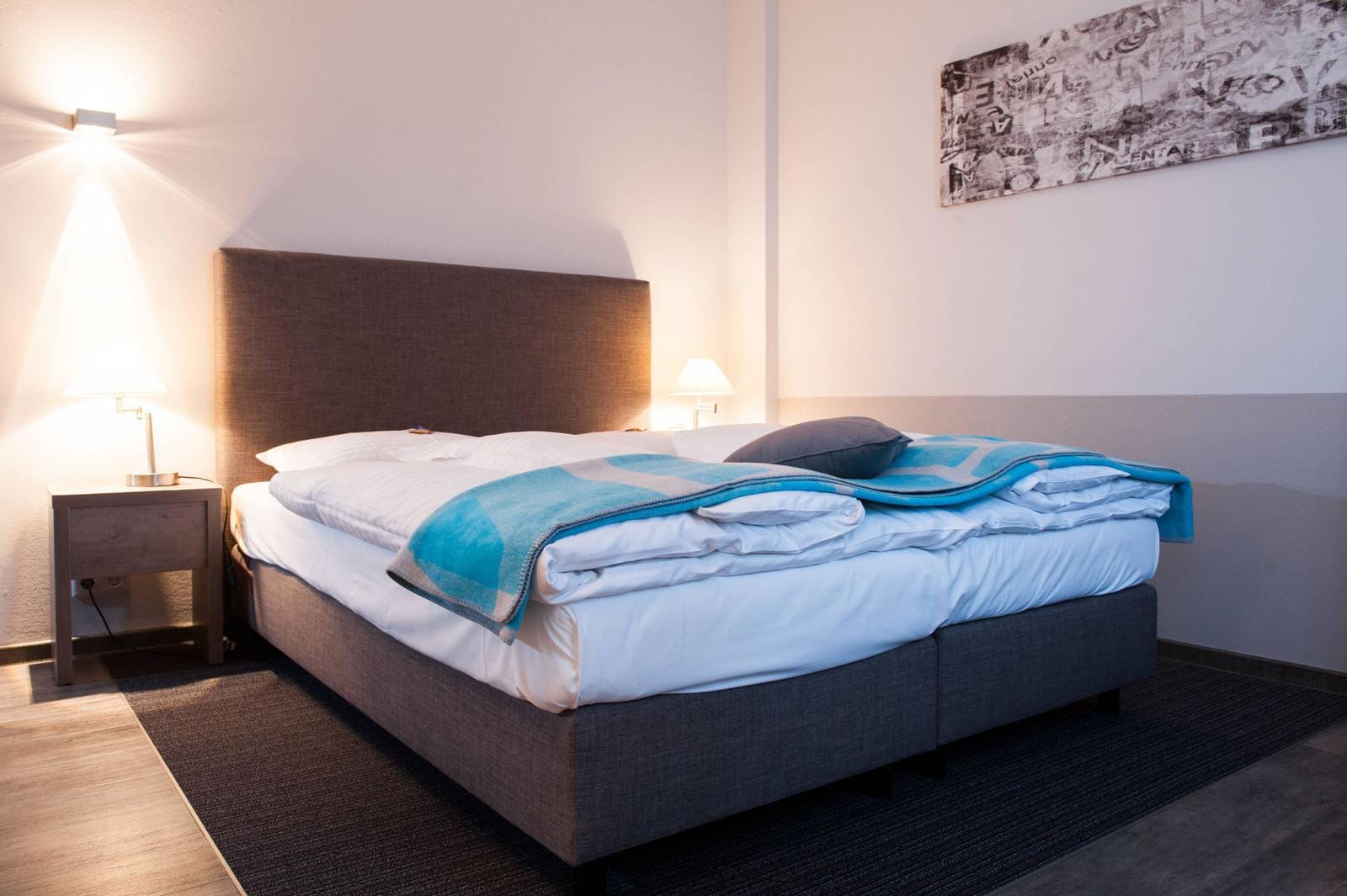 Modernes Hotelzimmer mit Doppelbett, graues Kopfteil, blaue Decke, Holzlampe, gemütlich.