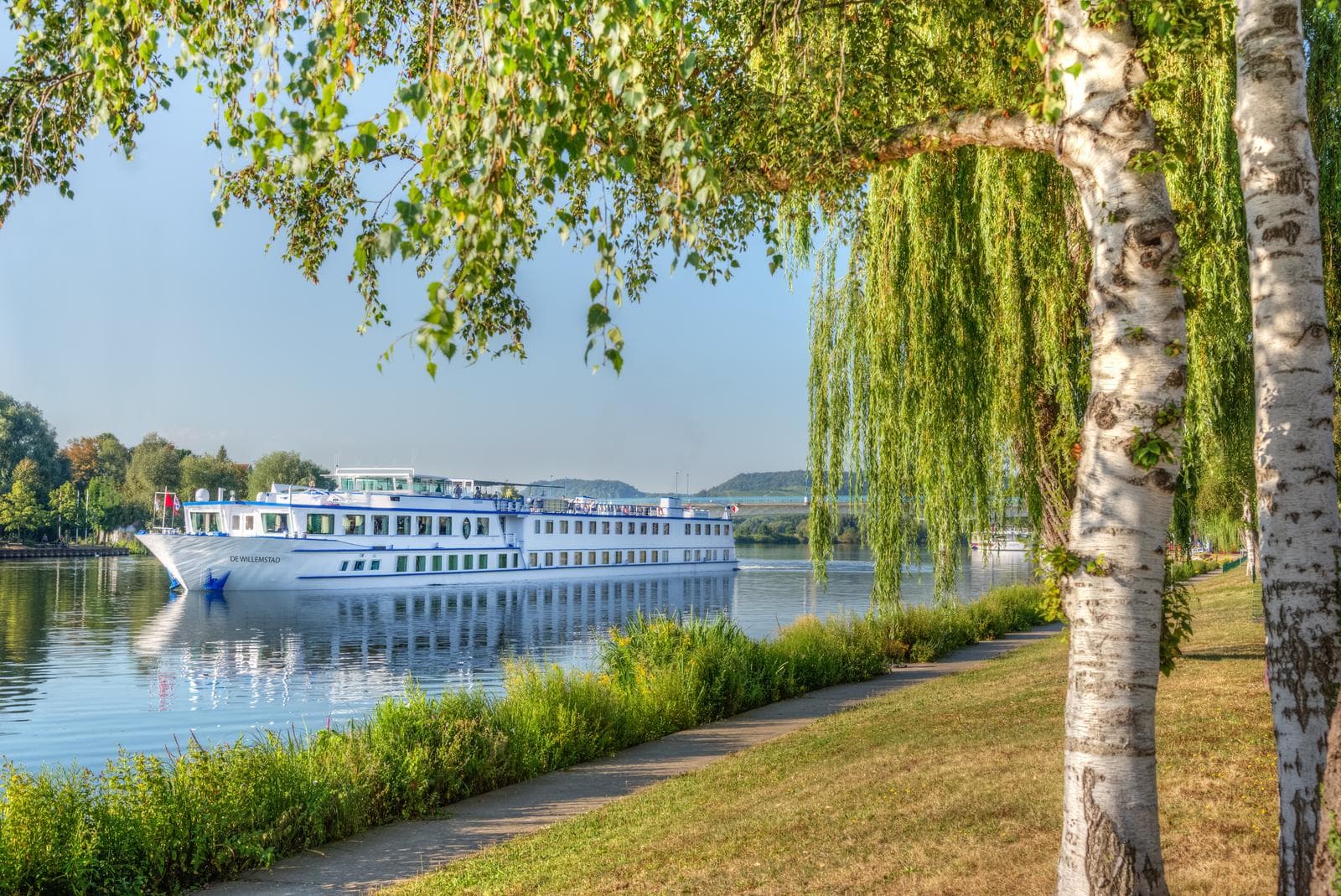 Ein weiĂes Flusskreuzfahrtschiff auf ruhigem Fluss, umgeben von grĂŒnen BĂ€umen.