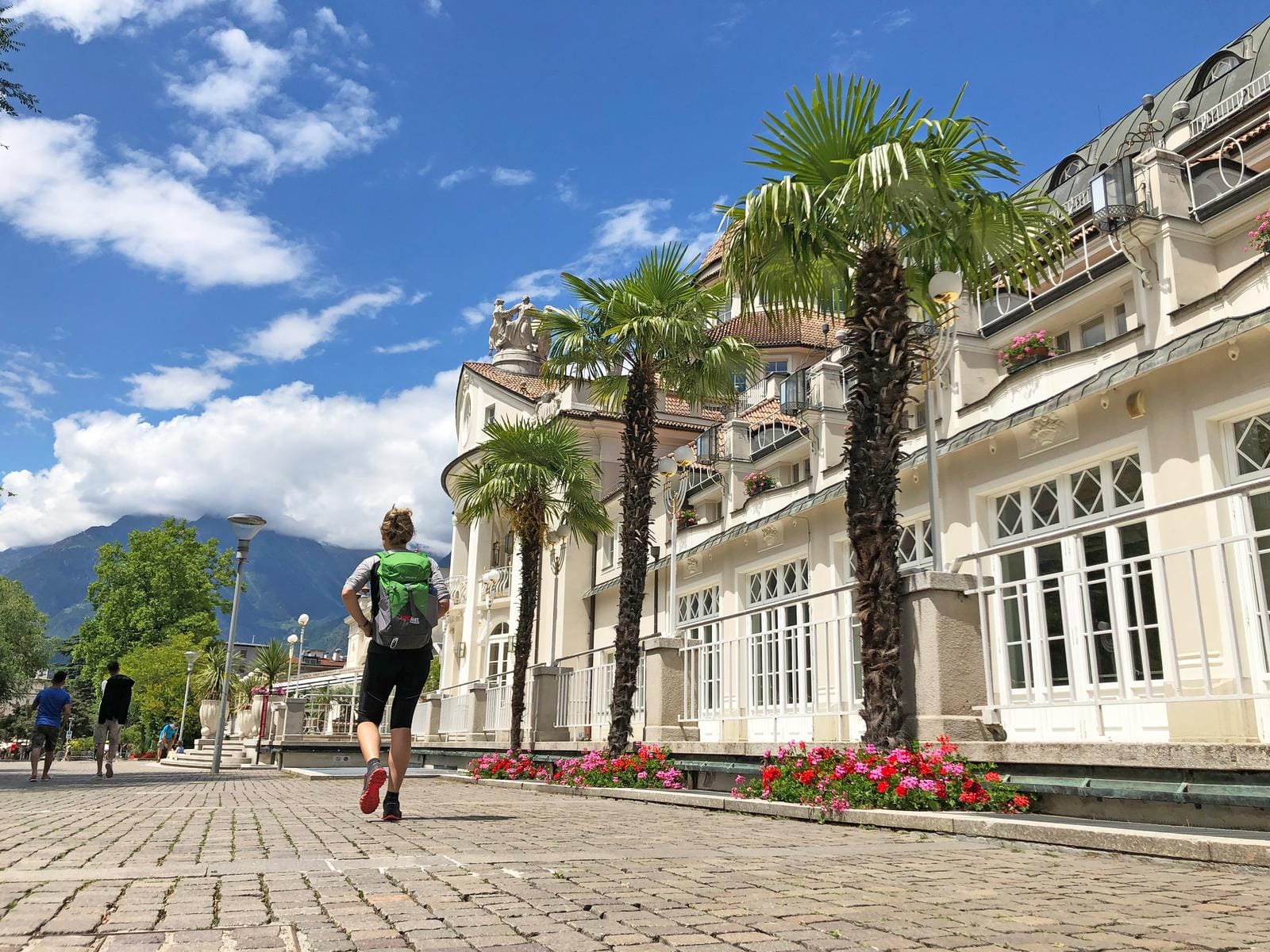 Promenade in Meran, Italien, mit Palmen, historischem Gebäude und Bergblick.