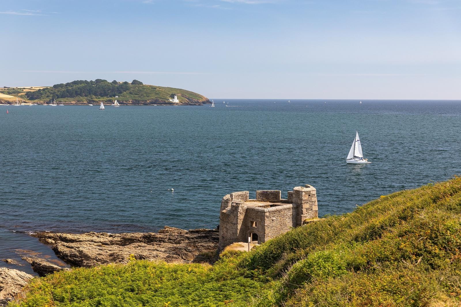 KĂĽste mit historischem Turm, Segelbooten und grĂĽnen HĂĽgeln in Cornwall, England.