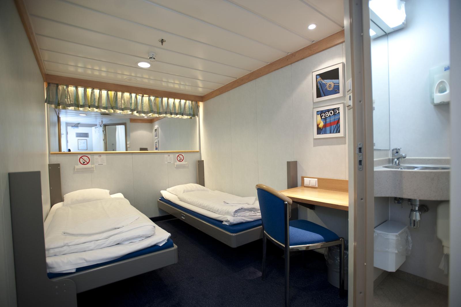 Zweibett-Schiffskabine mit Schreibtisch, blauem Teppich, hellem Holz und kleinem Bad.