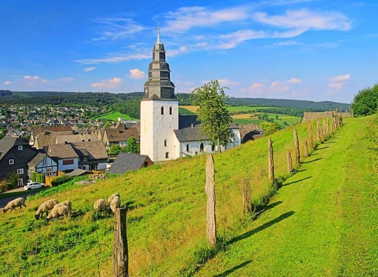 Landschaft mit Kirche und Schafen im Sauerland, Deutschland, unter blauem Himmel.