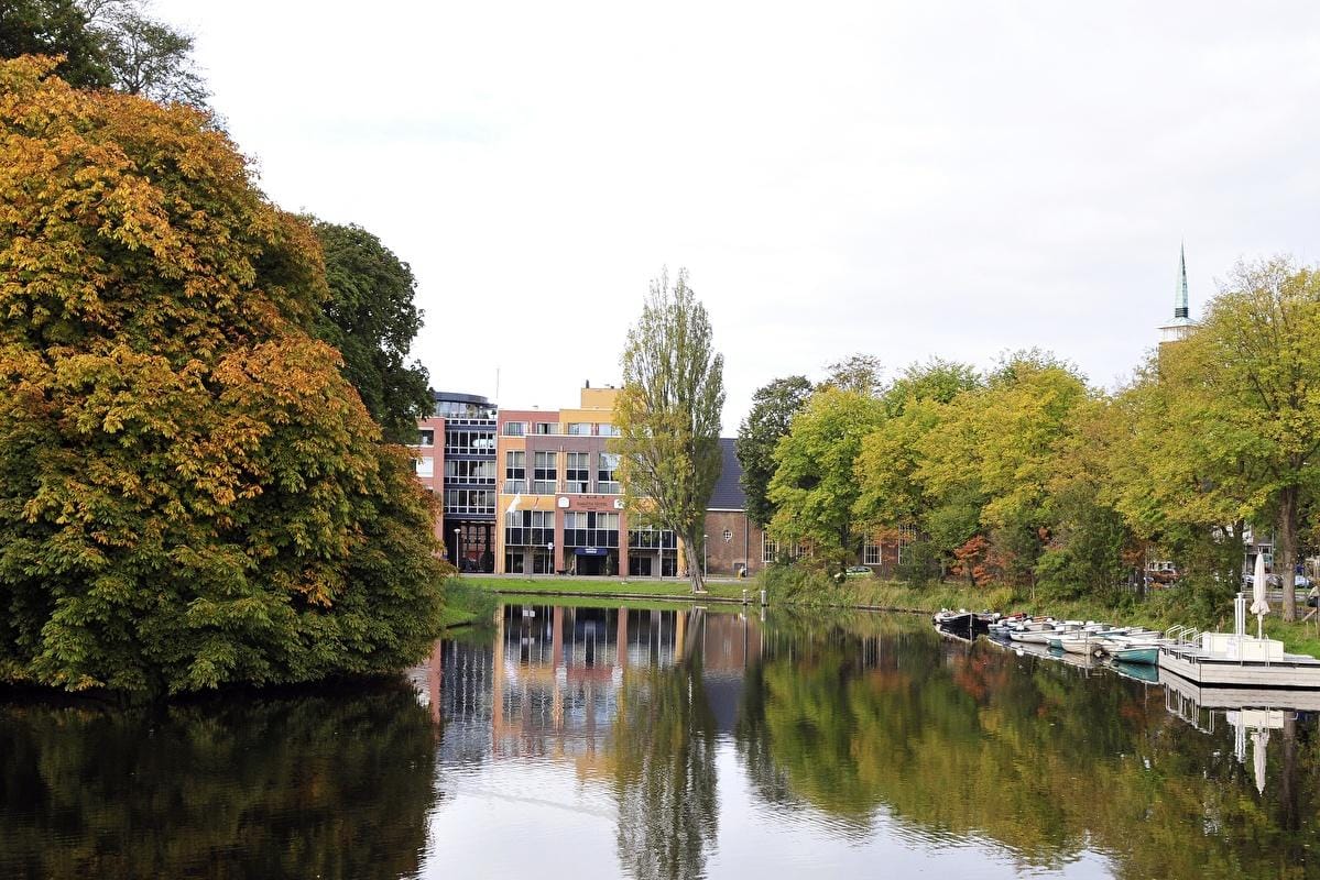 Herbstliche Landschaft mit See, bunten Bäumen, Booten und modernen Gebäuden.