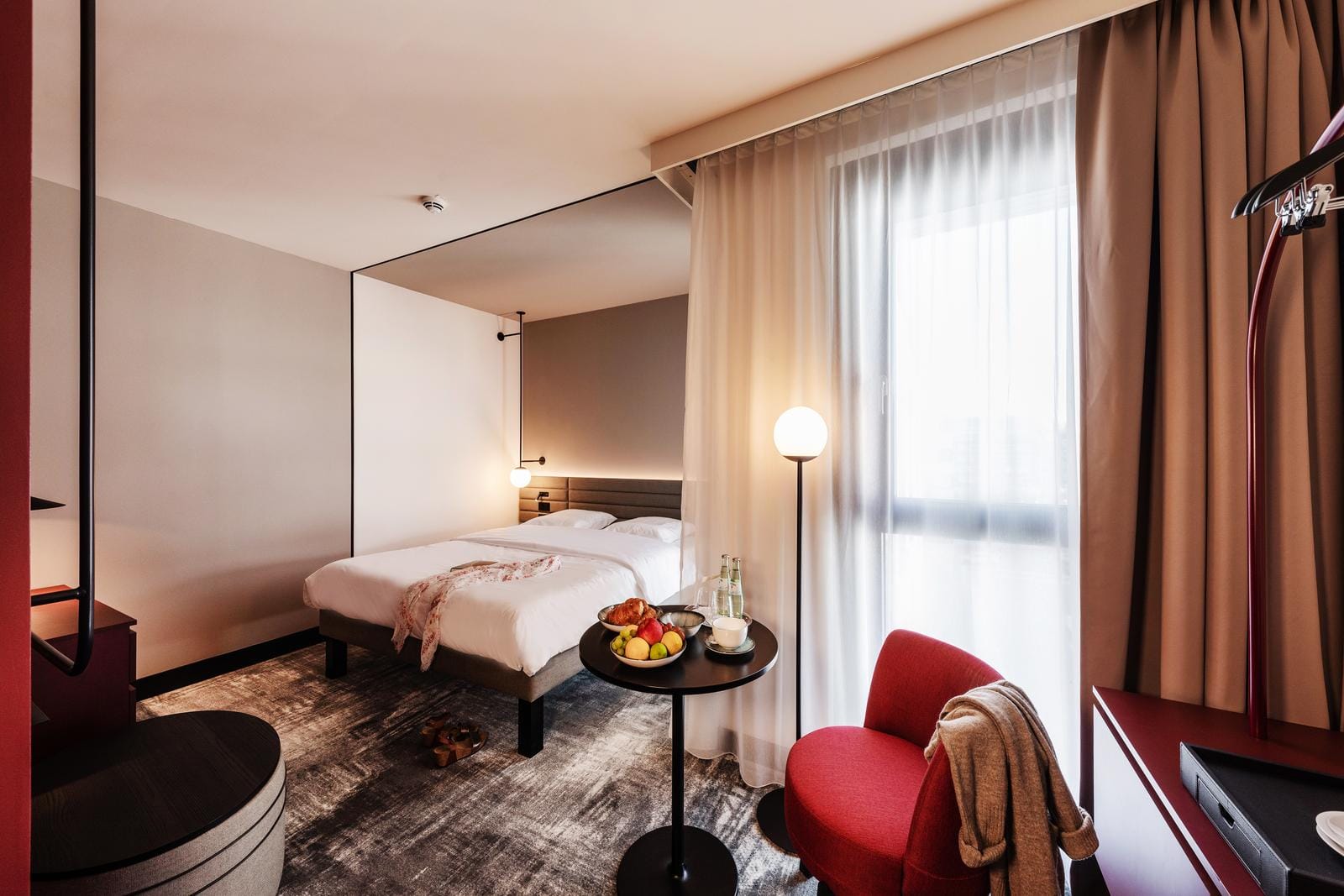 Modernes Hotelzimmer mit Doppelbett, rotem Sessel, Lampen und Obstteller.