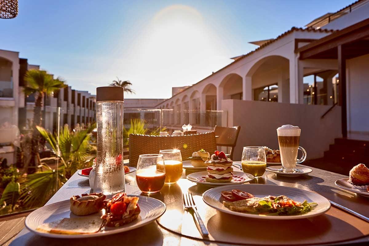 Frühstückstisch auf sonniger Hotelterrasse mit Palmen und mediterraner Architektur.