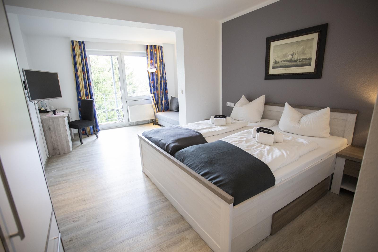 Modernes Hotelzimmer mit grauer Akzentwand, hellem Holzboden und großen Fenstern.