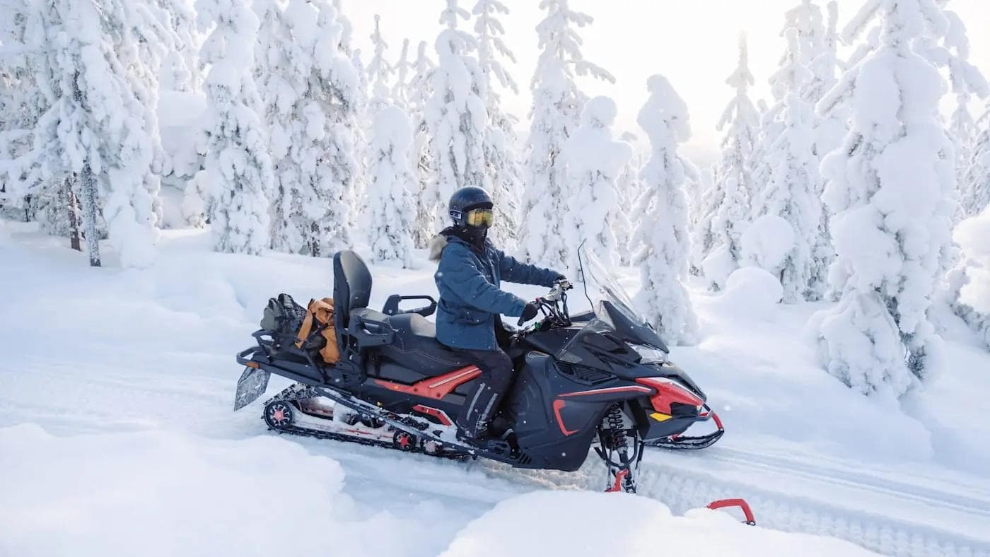 Person fährt Schneemobil durch verschneiten Wald in winterlicher Landschaft.