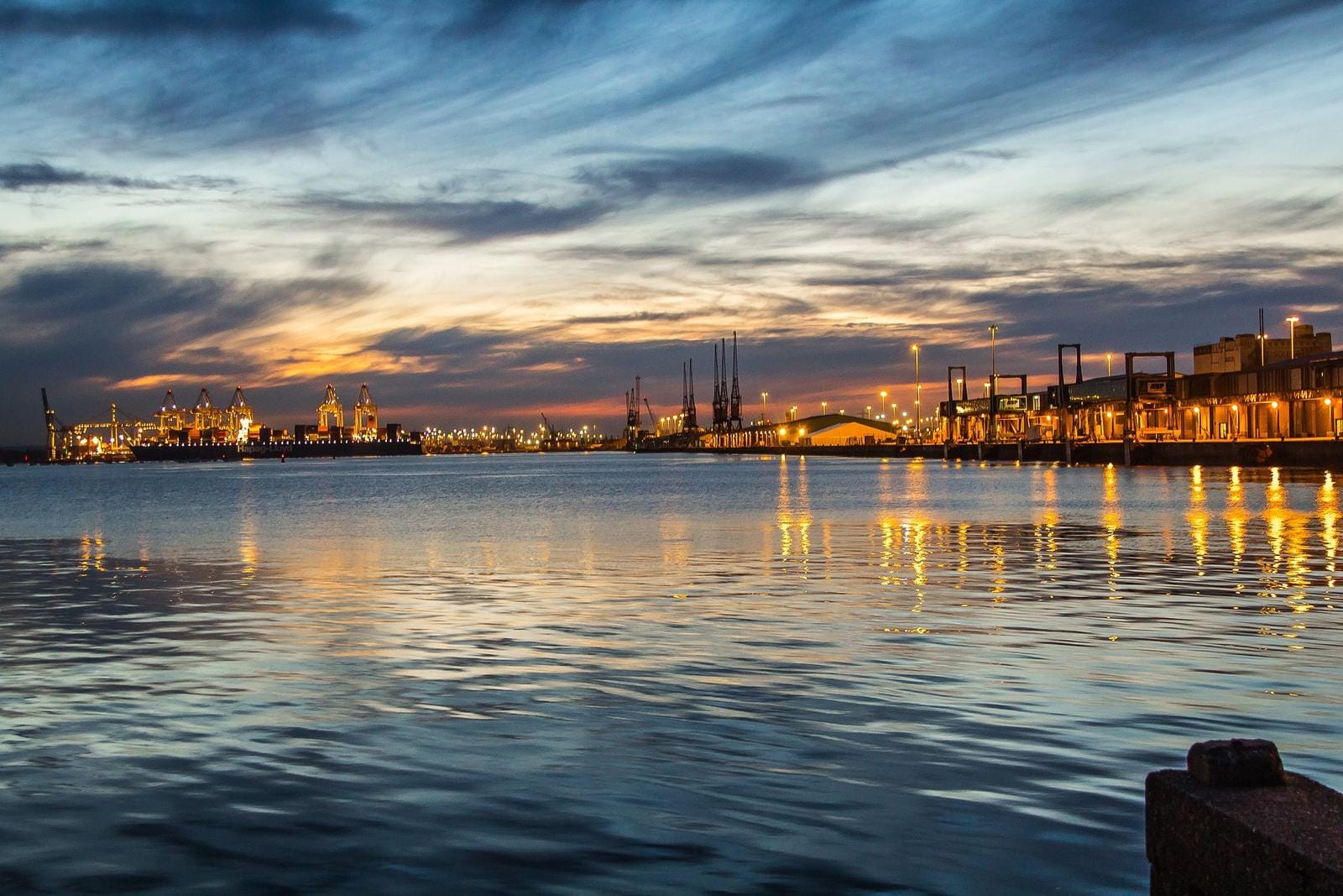 Hafenansicht bei Sonnenuntergang mit beleuchteten Kränen und reflektierendem Wasser.